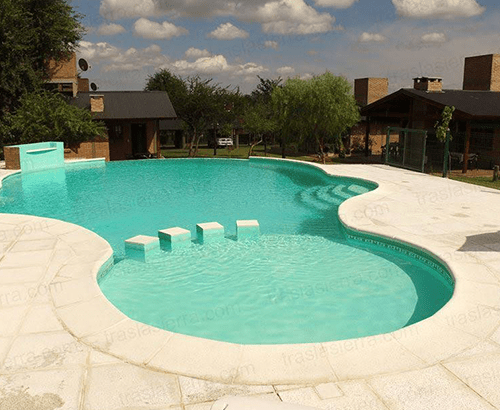 Piscina irregular con cascada y solárium húmedo circular para niños con desborde infinito. Losetas atérmicas de color blanco para espacio de estar y relajación, como así también de circulación alrededor de la piscina.